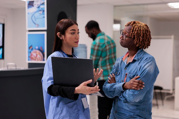 Mujer afroamericana y enfermera mirando una computadora portátil para encontrar un tratamiento de recuperación para el diagnóstico de atención médica en el vestíbulo de la instalación. Gente hablando de enfermedades y medicinas, usando computadoras en recepción.