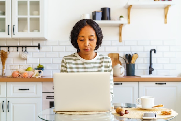 Foto mujer afroamericana enfocada trabajando desde la cocina en una computadora portátil concepto de oficina remota en el hogar