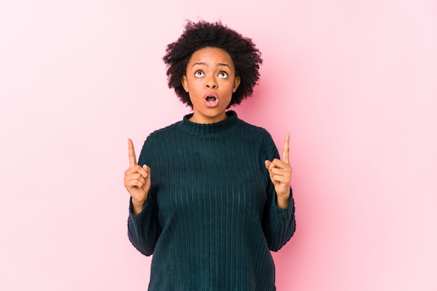 Mujer afroamericana envejecida media contra una pared rosada que señala al revés con la boca abierta.