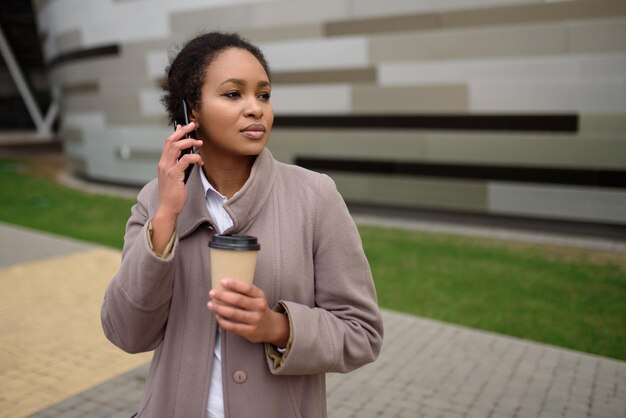 Una mujer afroamericana está hablando por un teléfono móvil y sosteniendo un café.