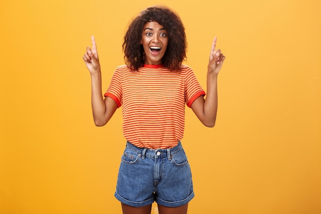 Mujer afroamericana con estilo levantando las manos y apuntando hacia arriba con una amplia sonrisa sobre la pared naranja