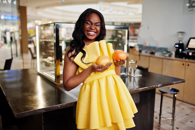 Mujer afroamericana con estilo en vestido amarillo posó en la cafetería con dos pomelos en las manos