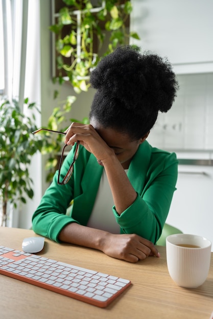 Una mujer afroamericana exhausta con dolor de cabeza se sienta en el escritorio de la oficina con el teclado Burnout