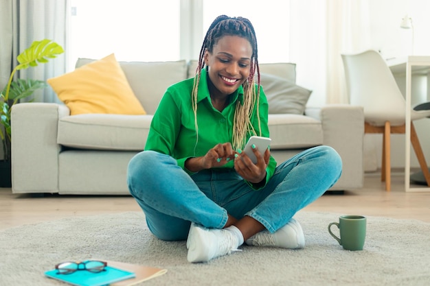 Mujer afroamericana feliz que usa un teléfono inteligente y una nueva aplicación móvil que envía mensajes de texto con amigos sentados en el piso en casa