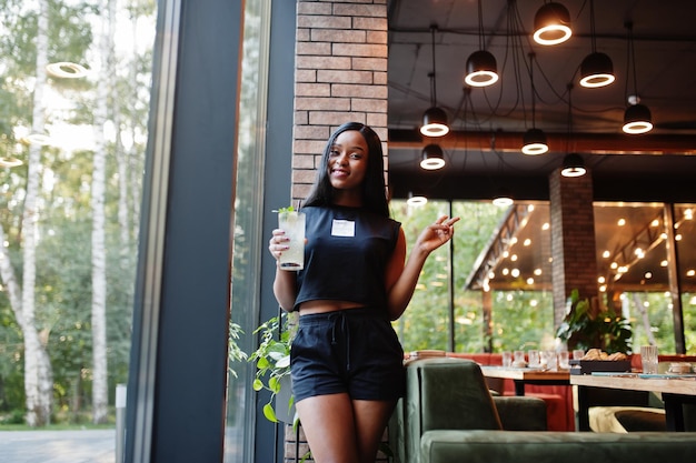 Mujer afroamericana feminista de moda vestida con camiseta negra y pantalones cortos, posada en un restaurante con vaso de limonada.