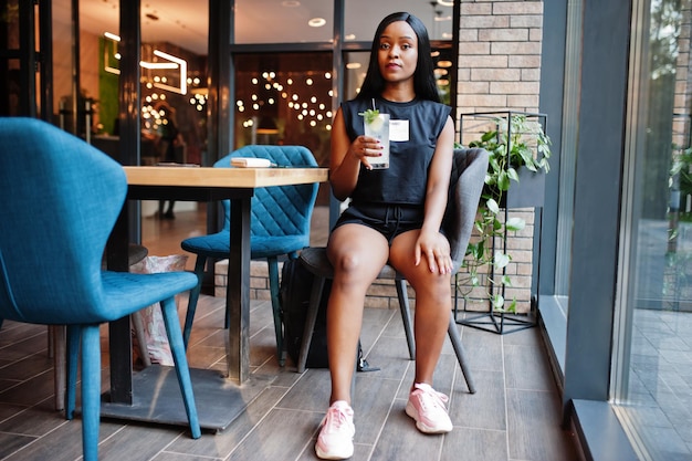 Mujer afroamericana feminista de moda vestida con camiseta negra y pantalones cortos, posada en un restaurante con vaso de limonada.