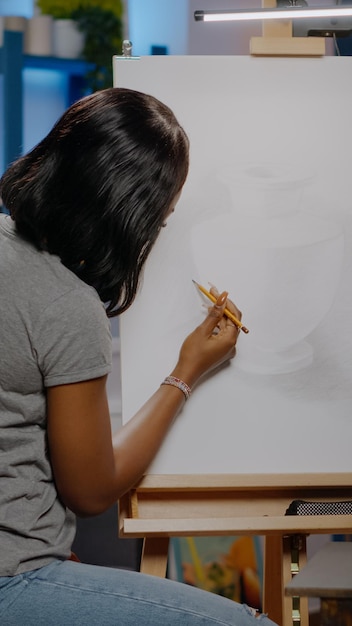 Mujer afroamericana con jarrón de dibujo de ocupación de artista sobre lienzo blanco con lápiz mientras está sentado en el estudio de creatividad. Artista joven negro trabajando en obra maestra de arte como pasatiempo artístico