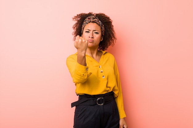 Mujer afroamericana joven contra un fondo rosado que muestra el puño a la cámara, expresión facial agresiva.