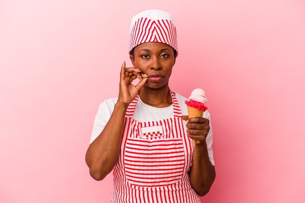 Mujer afroamericana joven fabricante de helado sosteniendo helado aislado sobre fondo rosa con los dedos en los labios guardando un secreto.