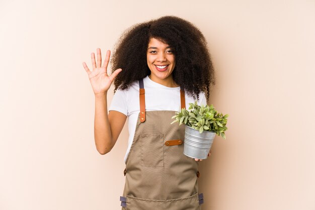 La mujer afroamericana joven del jardinero que sostenía una planta aisló sonreír alegre mostrando el número cinco con los dedos.