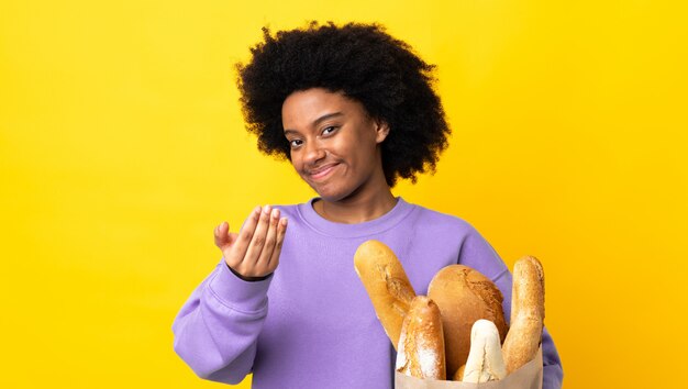 Mujer afroamericana joven que compra algo pan en la pared amarilla que invita a venir con la mano. Feliz de que hayas venido