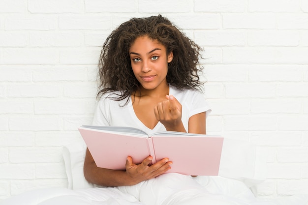 La mujer afroamericana joven que se sienta en la cama que estudia señalando con el dedo en usted como invitando se acerca.