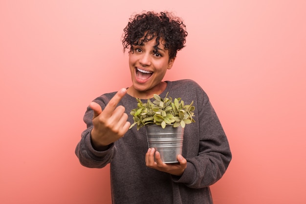 La mujer afroamericana joven que sostiene una planta que señala con el dedo en usted como invitando se acerca.