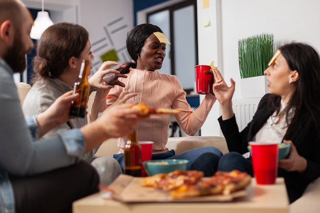 Mujer afroamericana jugando al juego de adivinanzas con amigos después del trabajo en la oficina. Grupo multiétnico de trabajadores adivinan la imitación para una actividad divertida y alegre mientras comen y beben cerveza