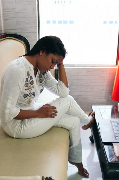 Mujer afroamericana llorando mientras está sentada en un sillón en el salón de su casa.