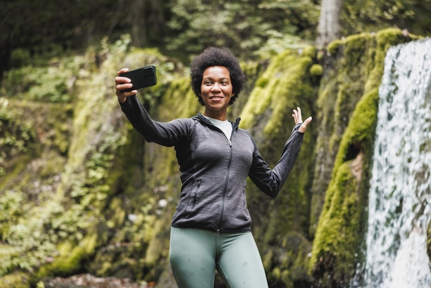 Mujer afroamericana madura haciendo una videollamada mientras está de pie cerca de la cascada y disfruta de la vista durante su caminata en las montañas.