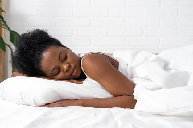 Mujer afroamericana con las manos debajo de la mejilla durmiendo en la cama con ropa de cama blanca