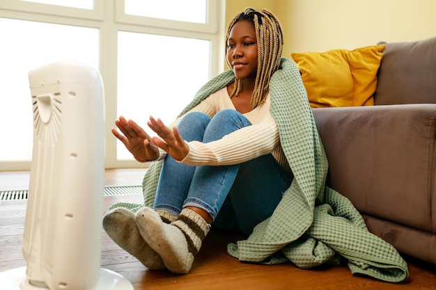 Mujer afroamericana en manta azul calentándose cerca del calentador eléctrico en casa