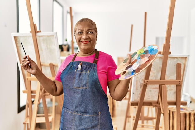 Mujer afroamericana mayor sonriendo confiada sosteniendo paleta y pincel en el estudio de arte