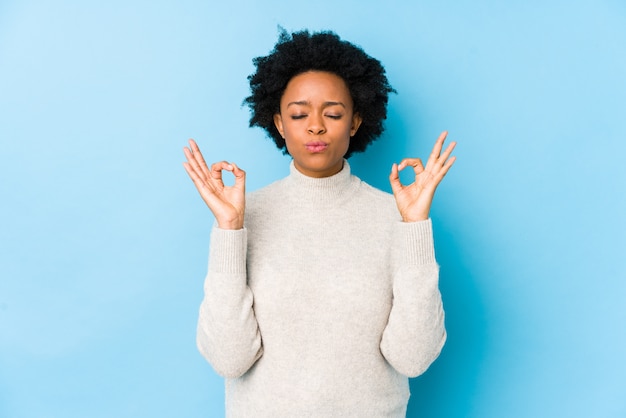 Mujer afroamericana de mediana edad contra una pared azul aislada se relaja después de un duro día de trabajo, ella está realizando yoga.