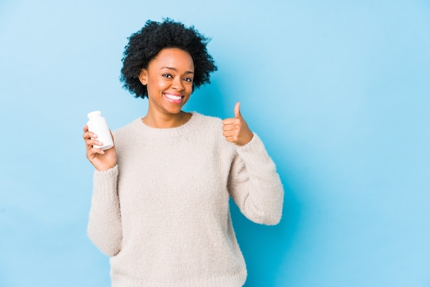 Mujer afroamericana de mediana edad sosteniendo una botella de vitamina sonriendo y levantando el pulgar hacia arriba