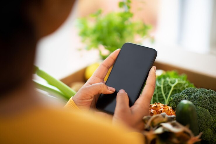Foto mujer afroamericana milenaria desempaqueta una caja de cartón con vegetales orgánicos usa un teléfono inteligente con pantalla en blanco