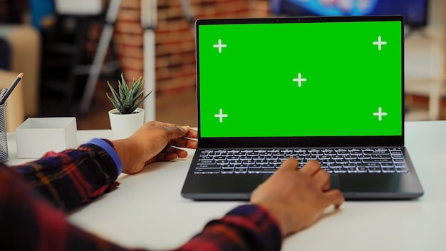 Mujer afroamericana mirando la pantalla verde en una computadora portátil en el escritorio, trabajando con una pantalla aislada y una plantilla de maqueta en una PC inalámbrica. Usando una computadora portátil con copyspace en blanco.