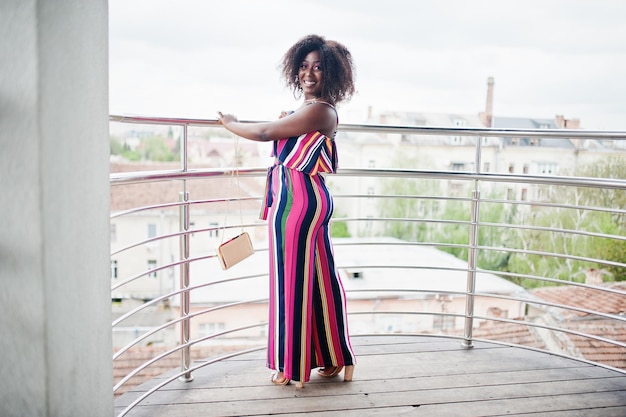 Mujer afroamericana de moda en mono rosa a rayas con bolso posado en el balcón francés