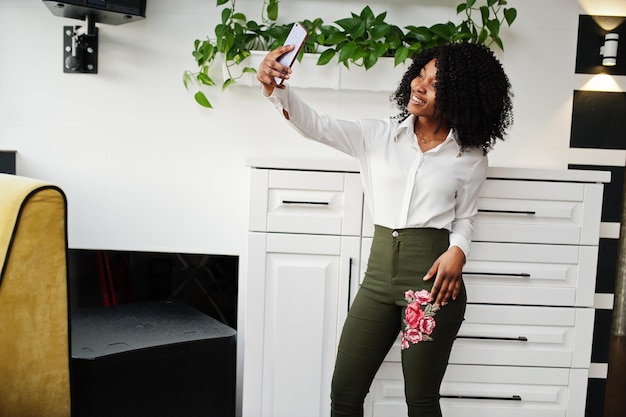 Mujer afroamericana de negocios pura con cabello afro, usa blusa blanca y pantalones verdes posada en un café con video en línea de transmisión de teléfono móvil