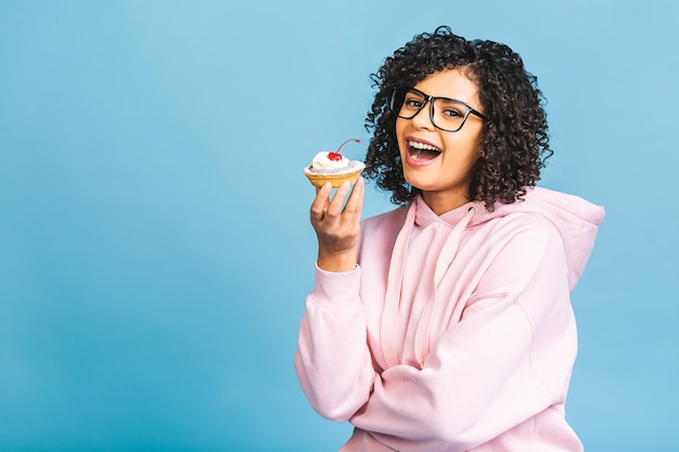 Mujer afroamericana negra feliz con peinado afro rizado haciendo un lío comiendo un enorme postre elegante sobre fondo azul. Comiendo cupcake.