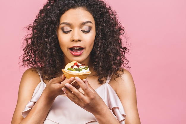 Mujer afroamericana negra feliz con peinado afro rizado haciendo un lío comiendo un enorme postre elegante sobre fondo rosa. Comiendo cupcake.