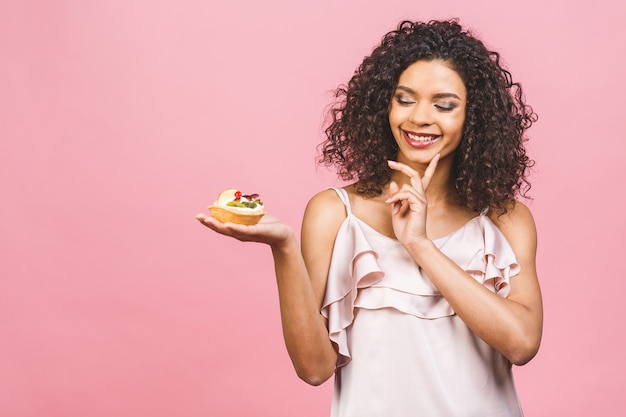 Mujer afroamericana negra feliz con peinado afro rizado haciendo un lío comiendo un enorme postre elegante sobre fondo rosa. Comiendo cupcake.