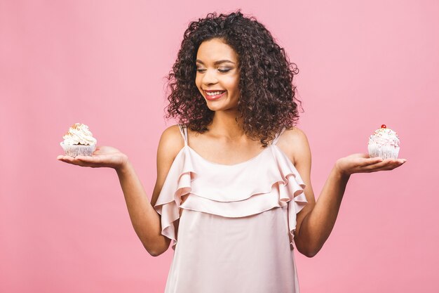 Mujer afroamericana negra feliz con peinado afro rizado haciendo un lío comiendo un enorme postre elegante sobre fondo rosa. Comiendo cupcake.