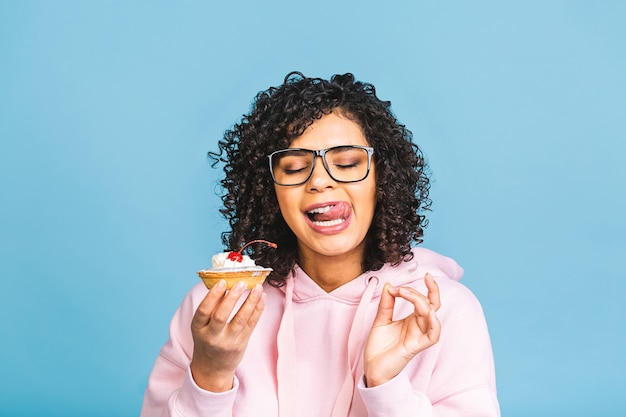 Mujer afroamericana negra feliz con peinado afro rizado haciendo un lío comiendo un postre de fantasía enorme sobre fondo azul. Comiendo cupcake.