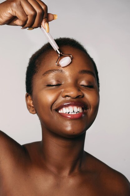 Mujer afroamericana con los ojos cerrados haciendo masaje facial con rodillo de jade aislado en la espalda blanca...