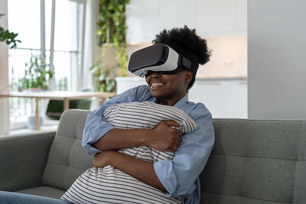 Mujer afroamericana optimista con auriculares VR sonriendo abrazando la almohada sintiendo miedo y deleite