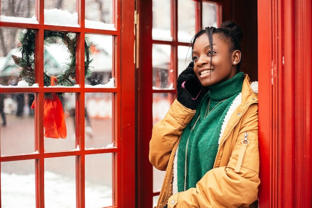 Mujer afroamericana parada en la calle cerca del mercado de la feria del árbol de Navidad, sostenga el teléfono parlante