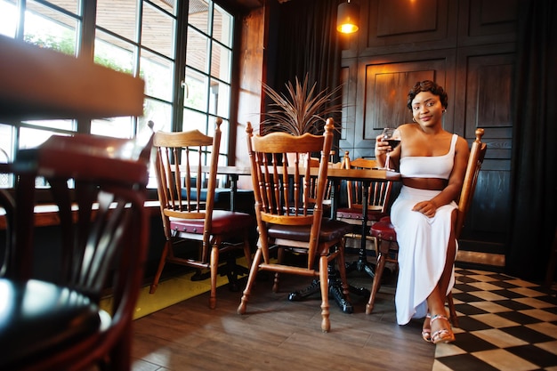 Mujer afroamericana, peinado retro con vestido blanco en el restaurante con copa de vino.