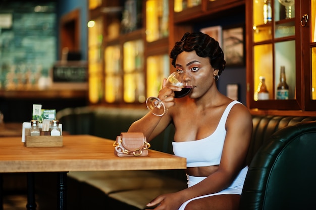 Mujer afroamericana, peinado retro con vestido blanco en el restaurante con copa de vino.