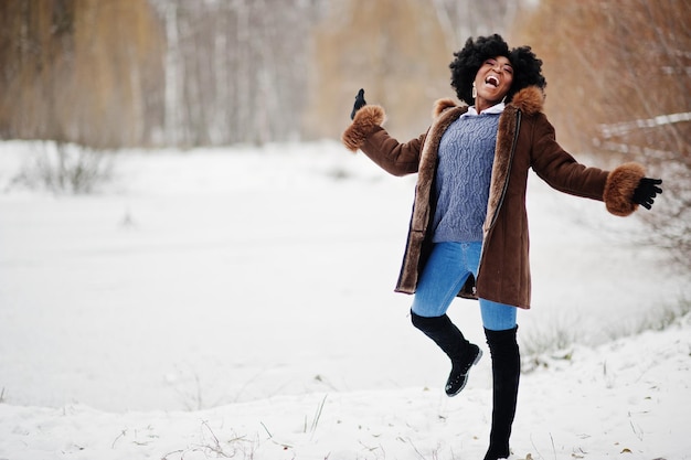 Mujer afroamericana de pelo rizado vestida con abrigo de piel de oveja y guantes posados en el día de invierno divirtiéndose y saltando