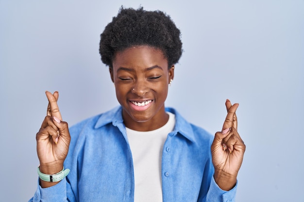 Mujer afroamericana de pie sobre fondo azul gesticulando con el dedo cruzado sonriendo con esperanza y ojos cerrados suerte y concepto supersticioso