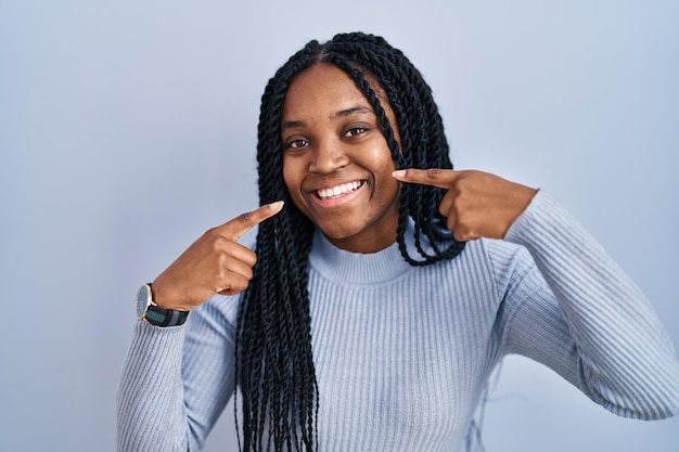 Mujer afroamericana de pie sobre fondo azul sonriendo alegre mostrando y señalando con los dedos los dientes y la boca concepto de salud dental