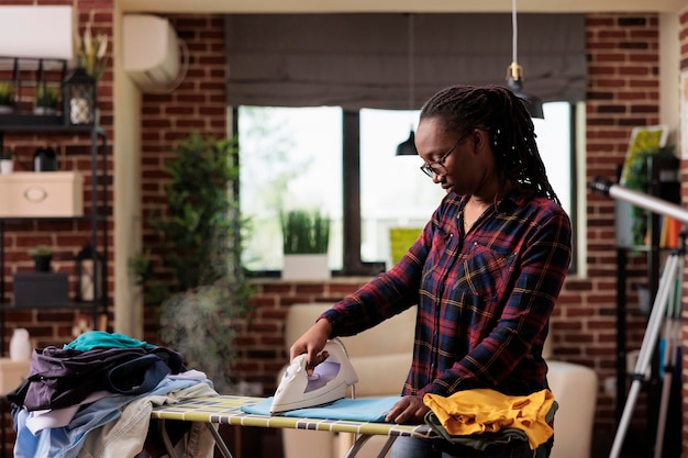 Mujer afroamericana planchando ropa en casa, día de lavado y planchado, estilo de vida independiente en apartamento urbano, ama de casa moderna haciendo tareas y tareas domésticas en casa. Vista frontal.