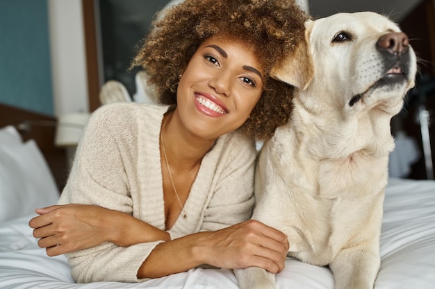 mujer afroamericana positiva con su labrador acostada en una cama en un hotel de viajes apto para mascotas