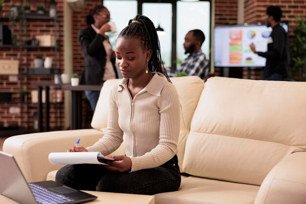 Mujer afroamericana que trabaja en archivos de informes para enviar correos electrónicos y planificar presentaciones comerciales en una computadora portátil. Gerente que usa documentos de papeleo e información de investigación para crear un proyecto corporativo.