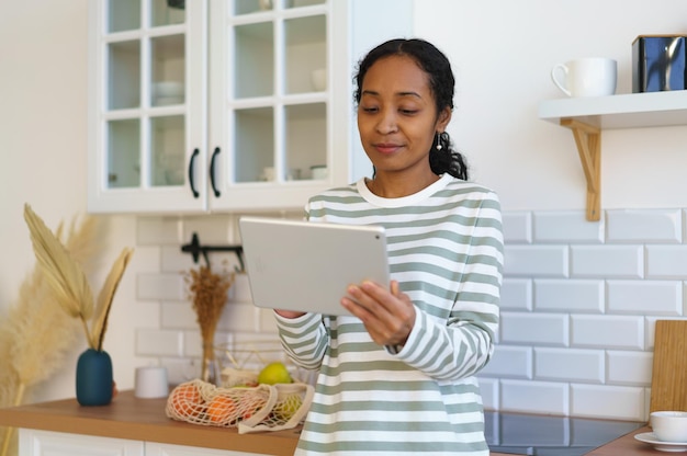 Mujer afroamericana que usa un dispositivo digital en la cocina buscando una receta antes de comenzar a cocinar