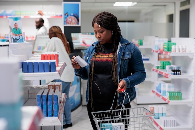 Mujer afroamericana revisando el tratamiento recetado en la farmacia, leyendo instrucciones sobre el paquete de vitaminas. Comprador eligiendo medicamentos, consultores de farmacia en segundo plano, todo el equipo negro