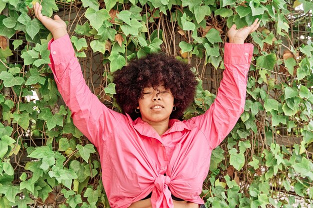 Mujer afroamericana segura de sí misma con los brazos levantados contra un fondo de vegetación mujer joven con cabello rizado y curvas