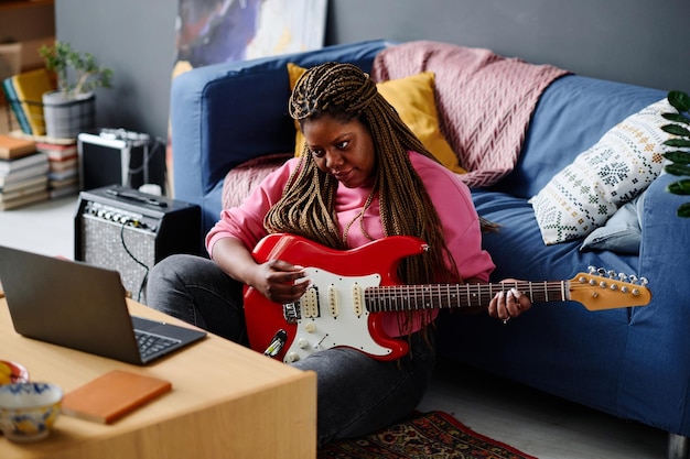 Mujer afroamericana sentada en el suelo frente a la computadora portátil y aprendiendo a tocar la guitarra en línea