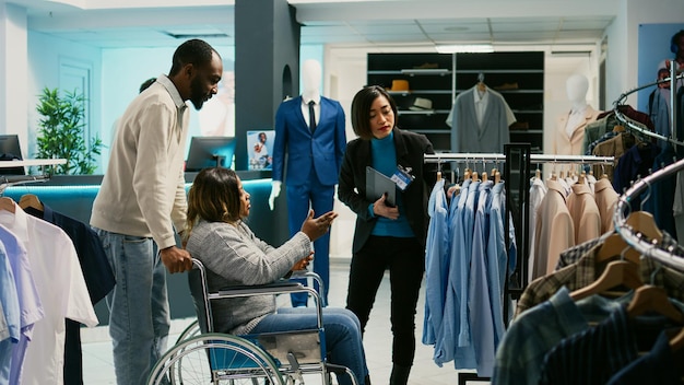 Mujer afroamericana en silla de ruedas comprando en un centro comercial, examinando la colección de ropa moderna en una tienda de ropa. Comprador masculino con discapacidad física comprando ropa informal, centro comercial.